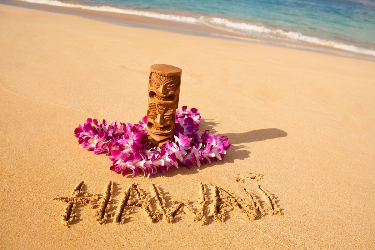 Word ‘Hawaii’ written in sand beside a tiki statue and lei on Big Island.